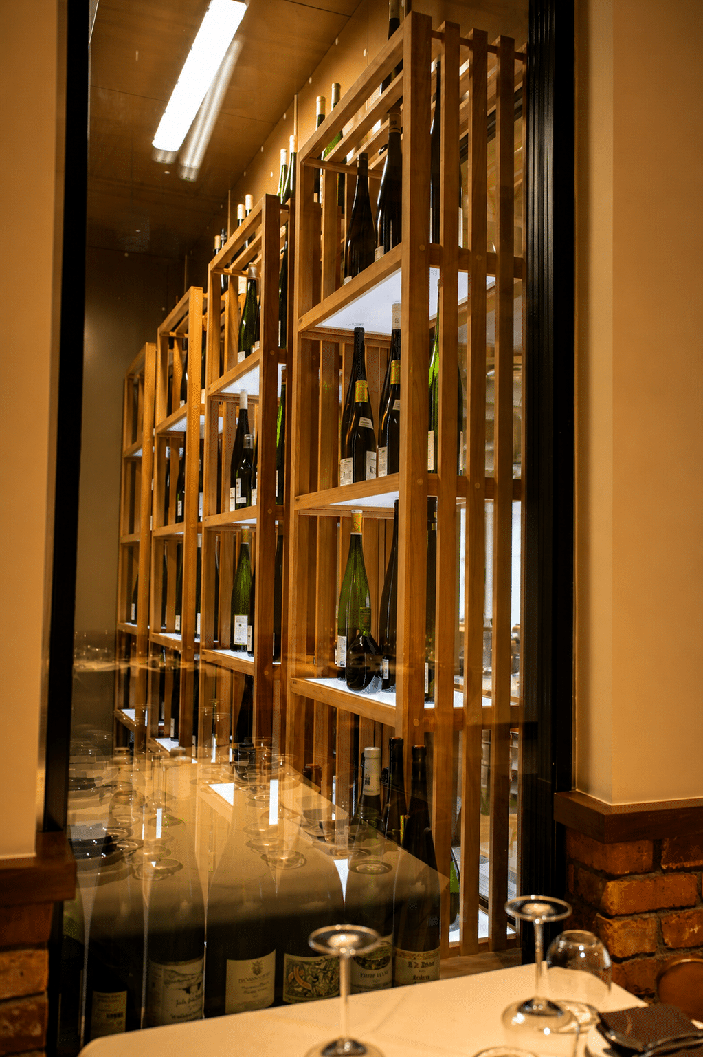 Wine bottles displayed on illuminated wooden shelves behind the restaurant wine room glass.